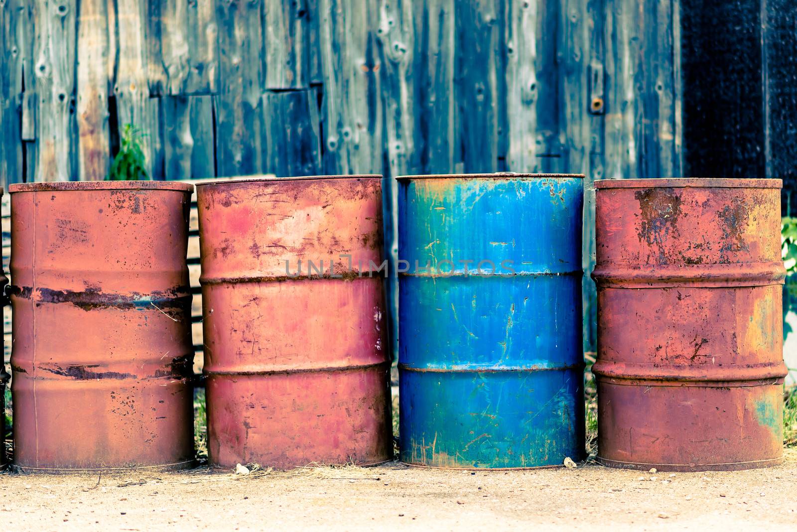 Photo presents four old used rust barrels for oil, petroleum, crude oil, mineral oil or petrol, three barrels are red and one is blue. In background visible wooden storehouse, warehouse.