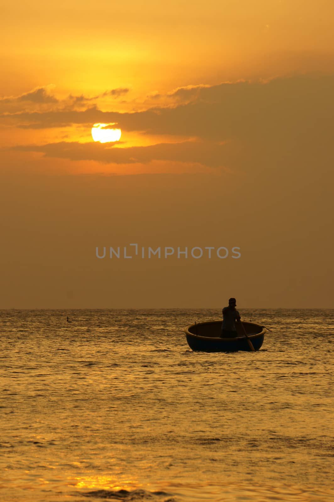 Beautiful landscape on ocean with silhouette of fisherman standing and rowing on circle boat at sunset, the sun go down, sky in brilliant yellow at sunset