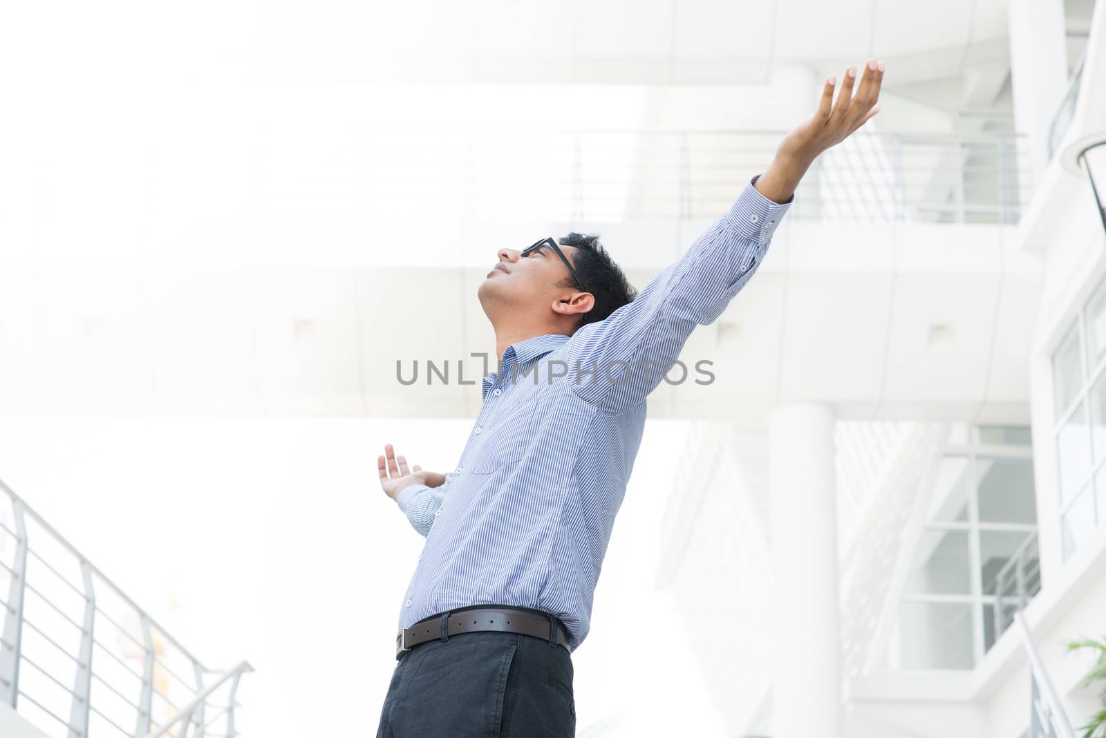 Successful Asian Indian businessman with arms outstretched celebrating his victory, modern office building as background, freedom concept.