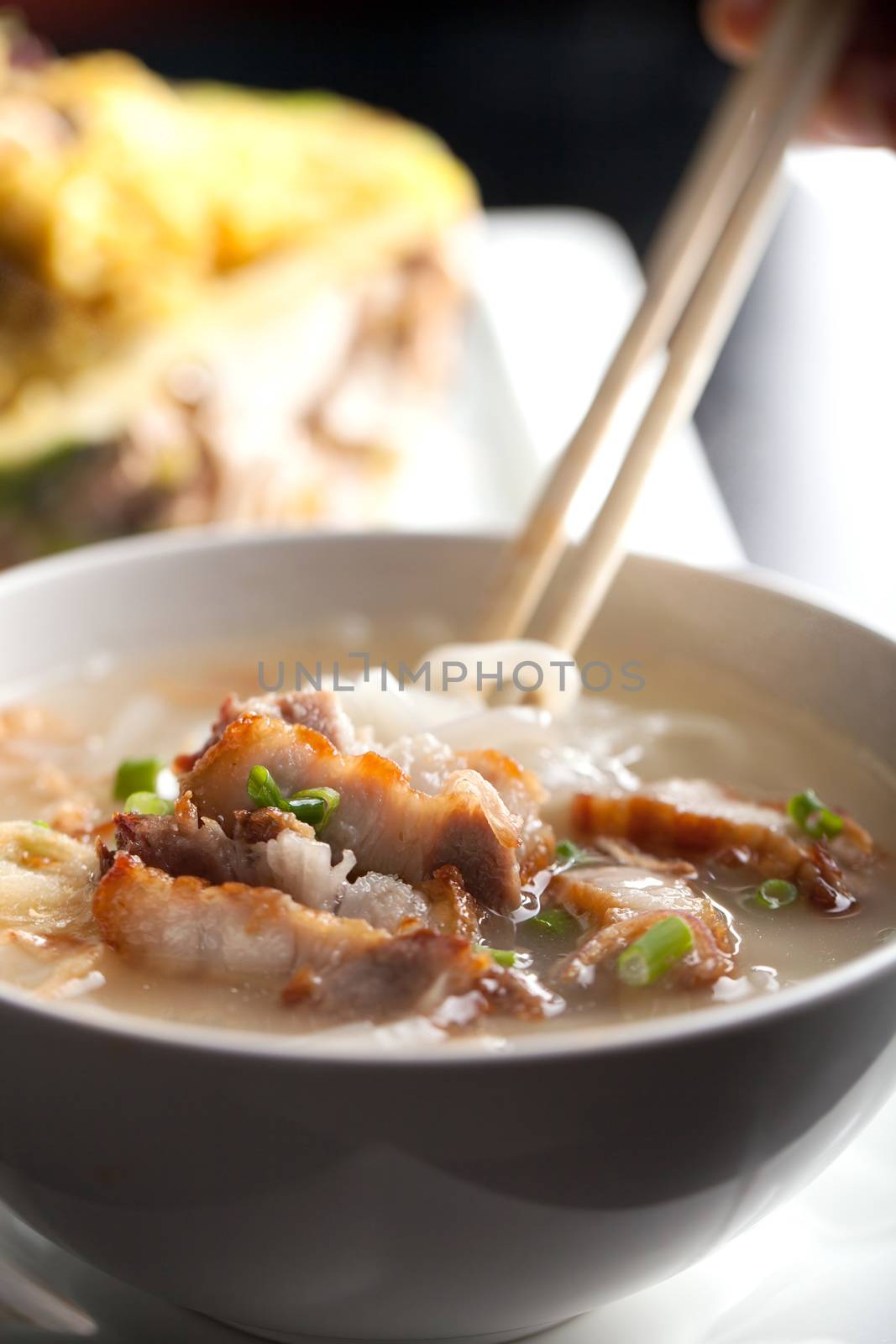 Closeup of a person eating Thai style crispy pork rice noodle soup from a bowl with chopsticks. Pineapple fried rice in the background.