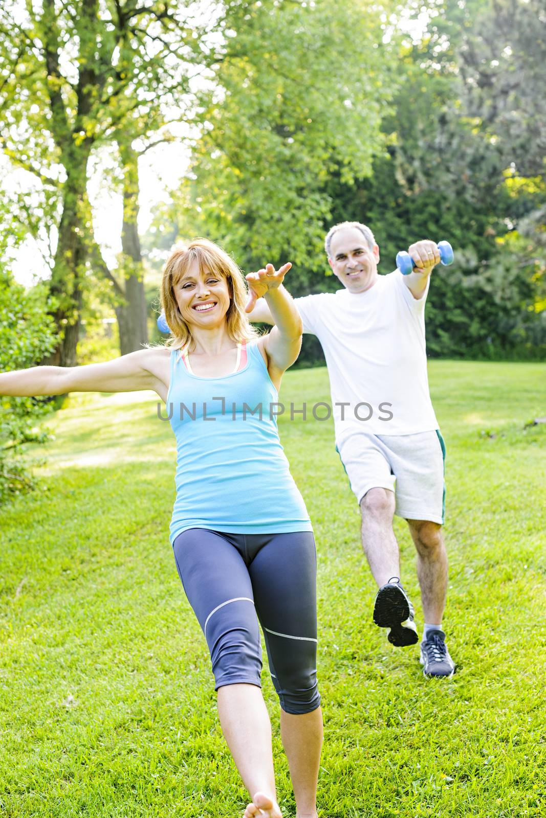 Female fitness instructor exercising with middle aged man outdoors in green park