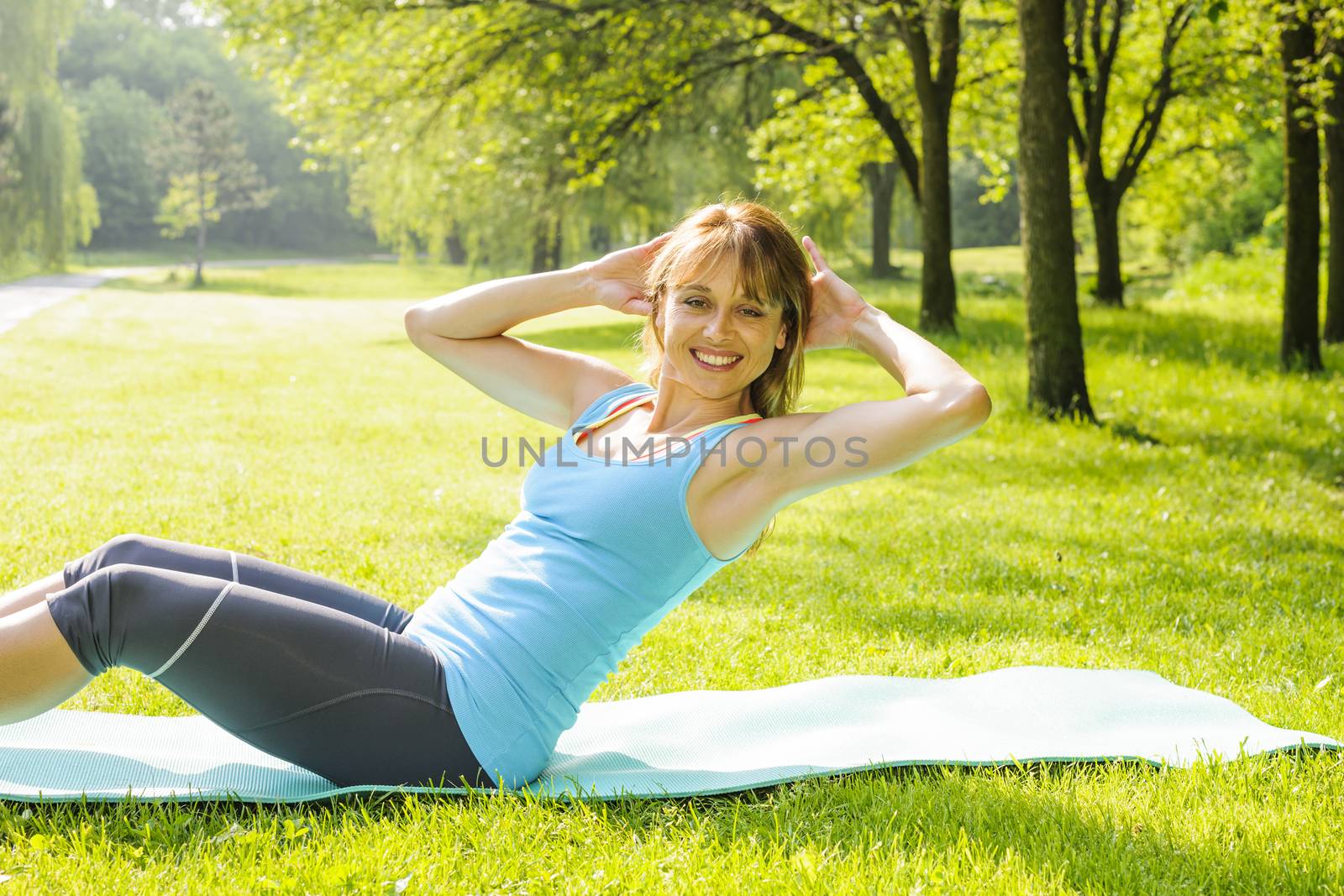 Female fitness instructor exercising doing sit-ups on yoga mat outside in green summer park