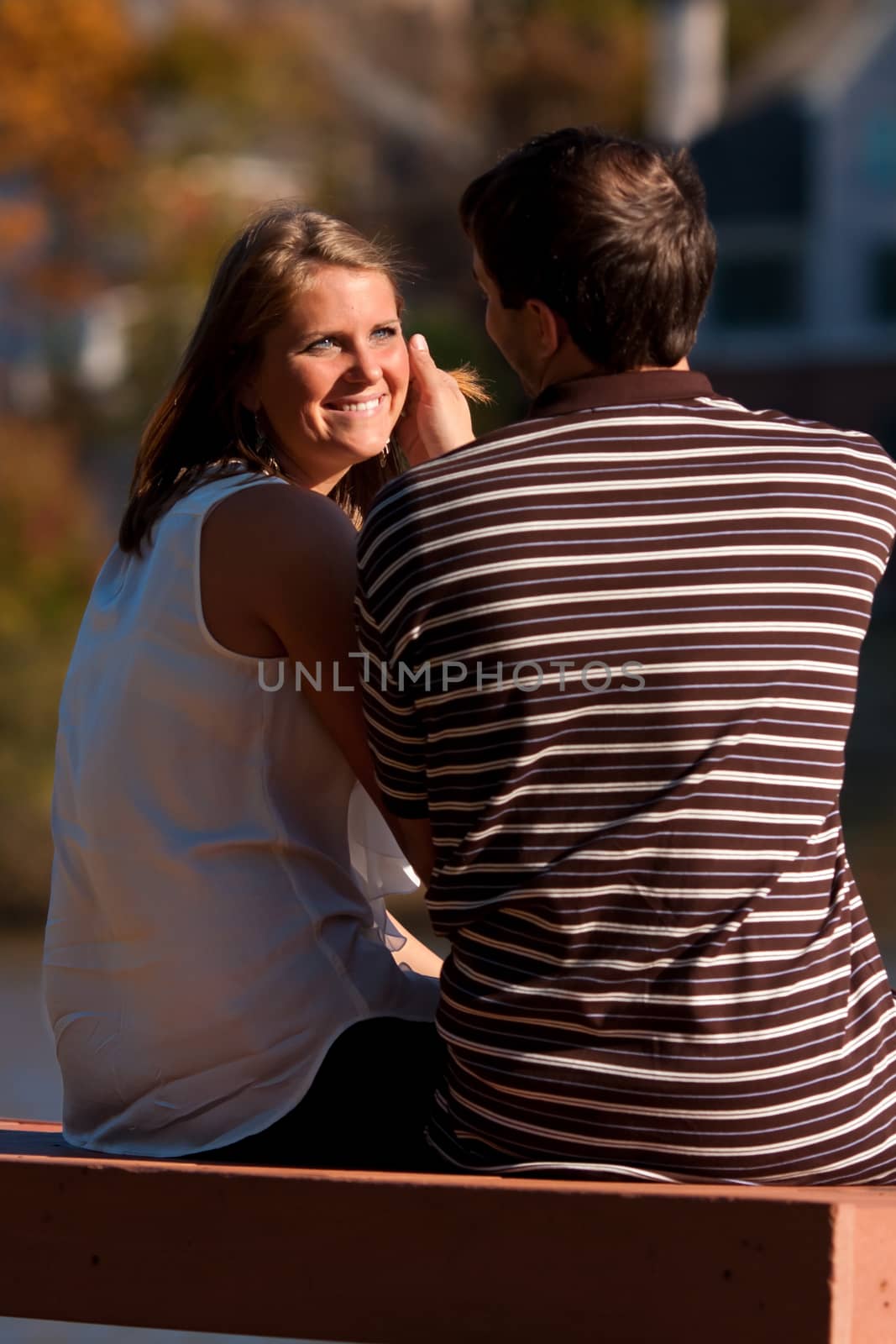 A young man gently caresses the smiling, sunlit face of his girlfriend while sitting on a bench.