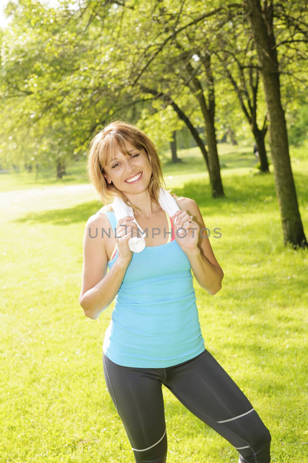 Smiling female fitness instructor standing with towel around neck after exercising in green park