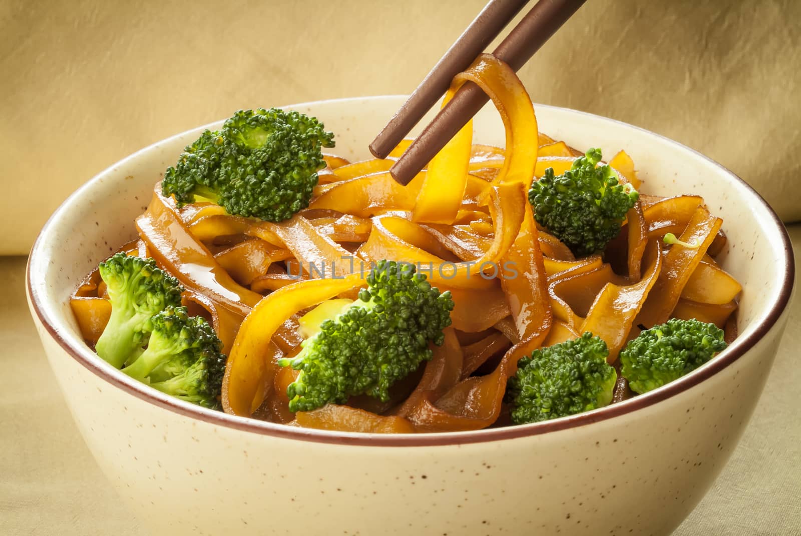 A bowl with delicious flat rice noodles and broccoli with chopsticks picking up a noodle.