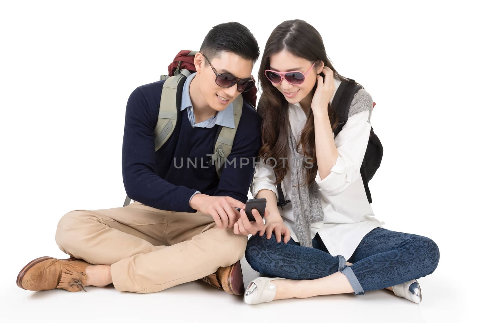 Happy traveling Asian couple sitting on ground and using cellphone, full length portrait on white background.