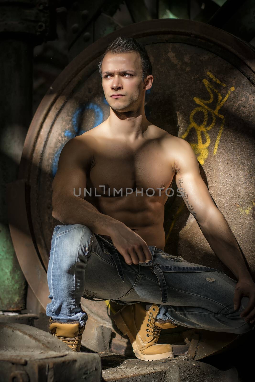 Muscular shirtless young man in abandoned industrial warehouse kneeling, looking away