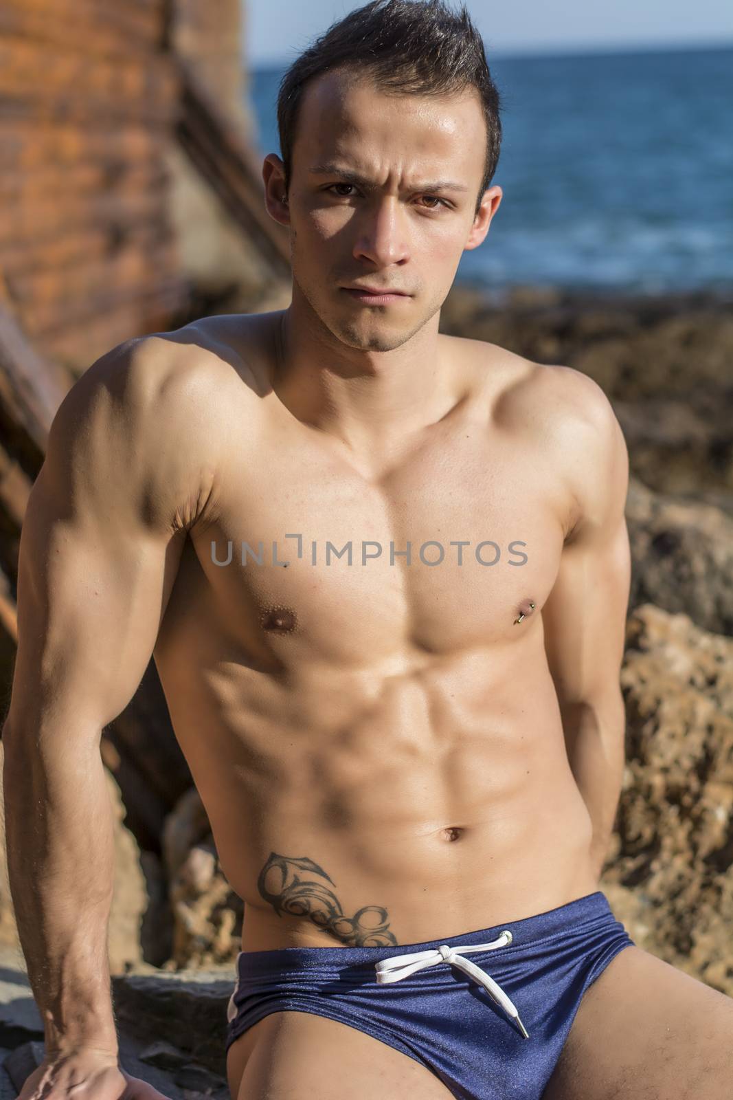 Muscular shirtless young man sunbathing on rock by the sea or ocean, looking at camera