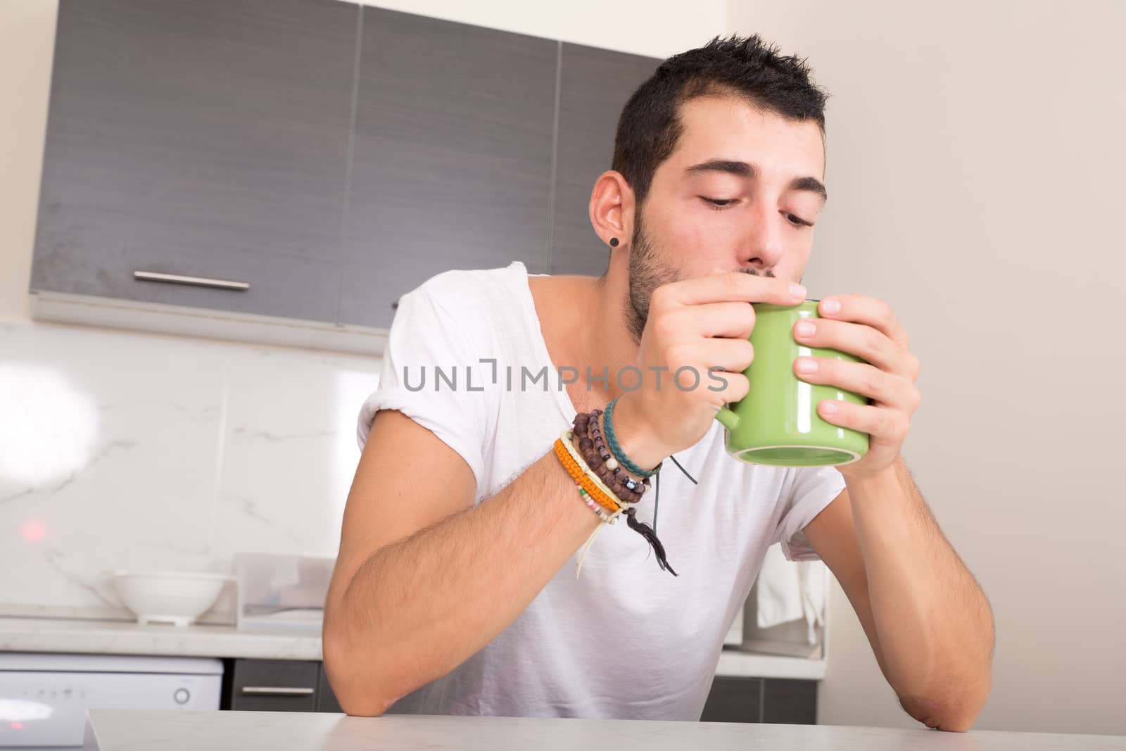 A young man sitting in the kitchen and drinking coffee.