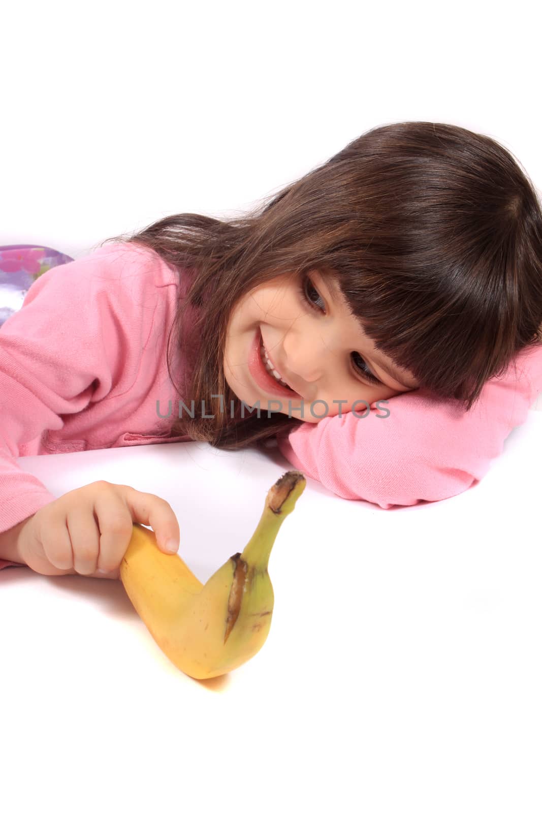 Young little girl laying on white background holding a banana and smiling