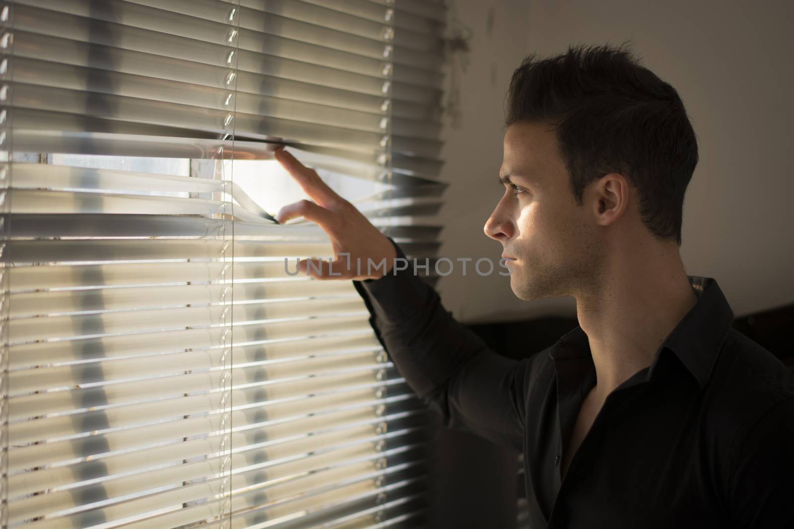 Profile of young man peeking through venetian blinds in a dark room