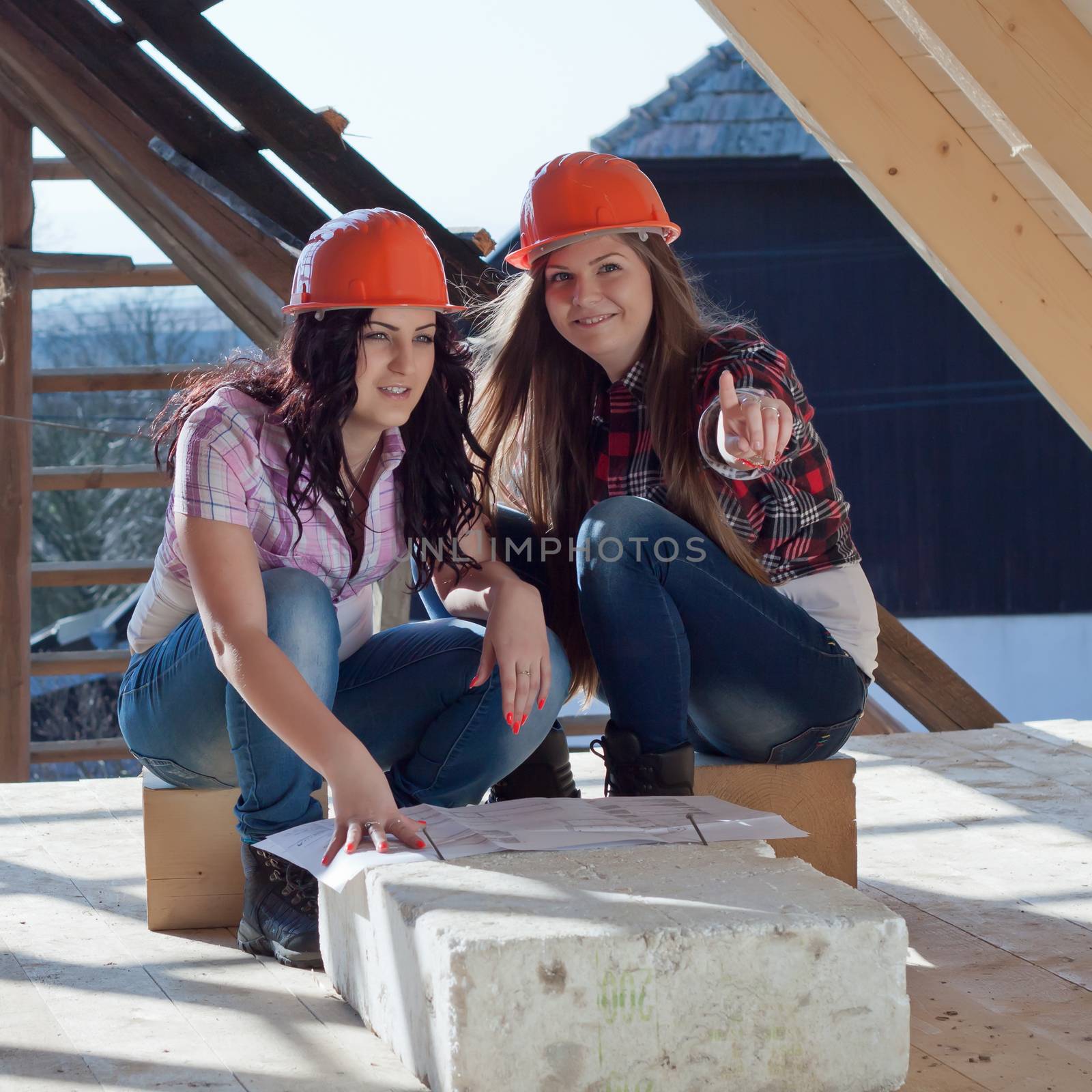 Two young women workers sitting on under construction roof, and control building in accordance with drawing
