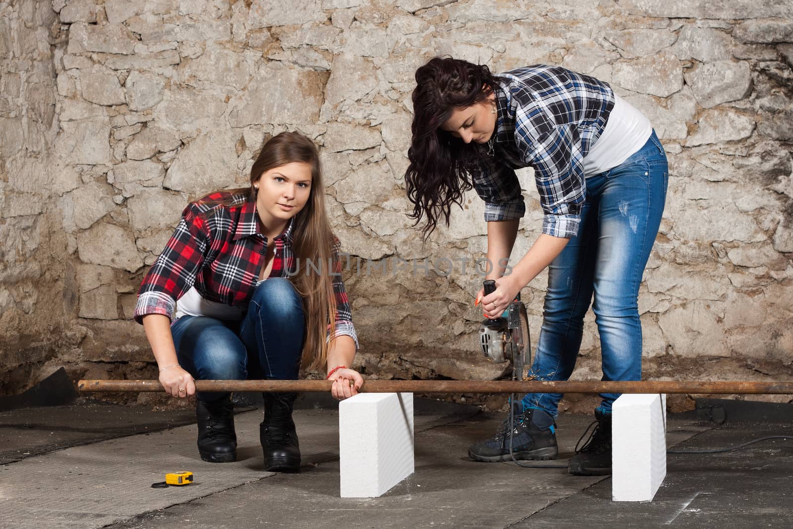 Two young long-haired woman cut iron pipe with angle grinder during the repair an old house