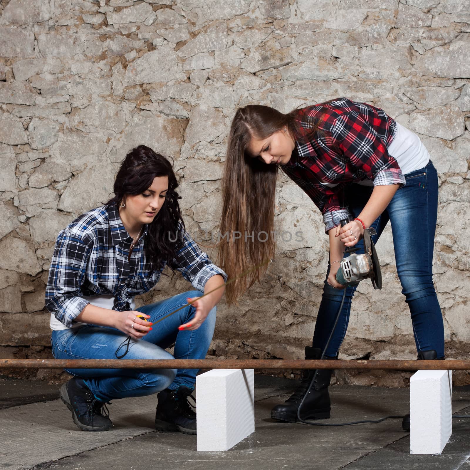 Two young long-haired woman cut iron pipe with angle grinder during the repair an old house