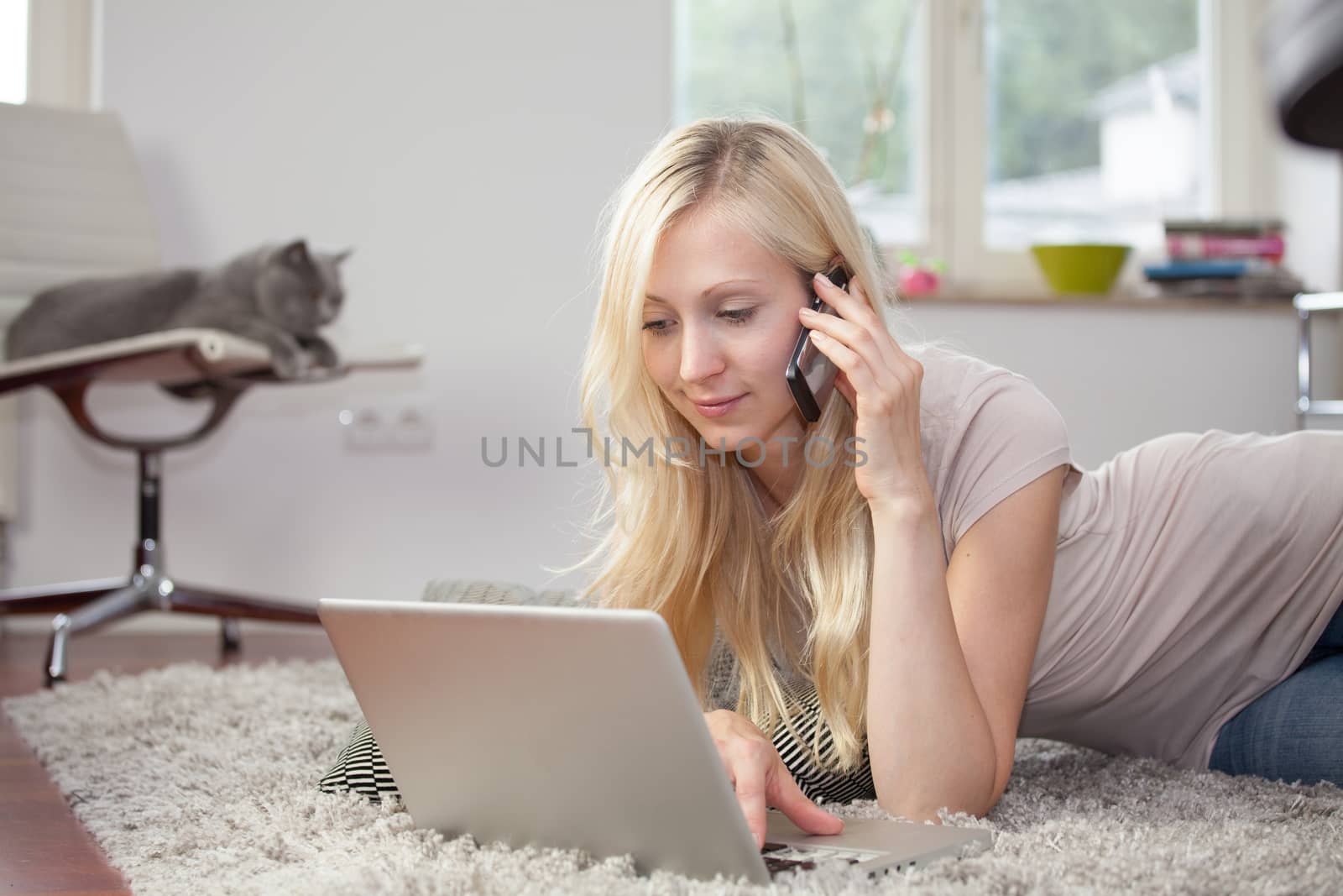 Relaxed woman is using the laptop and telephone in her livingroom, lying on the carpet