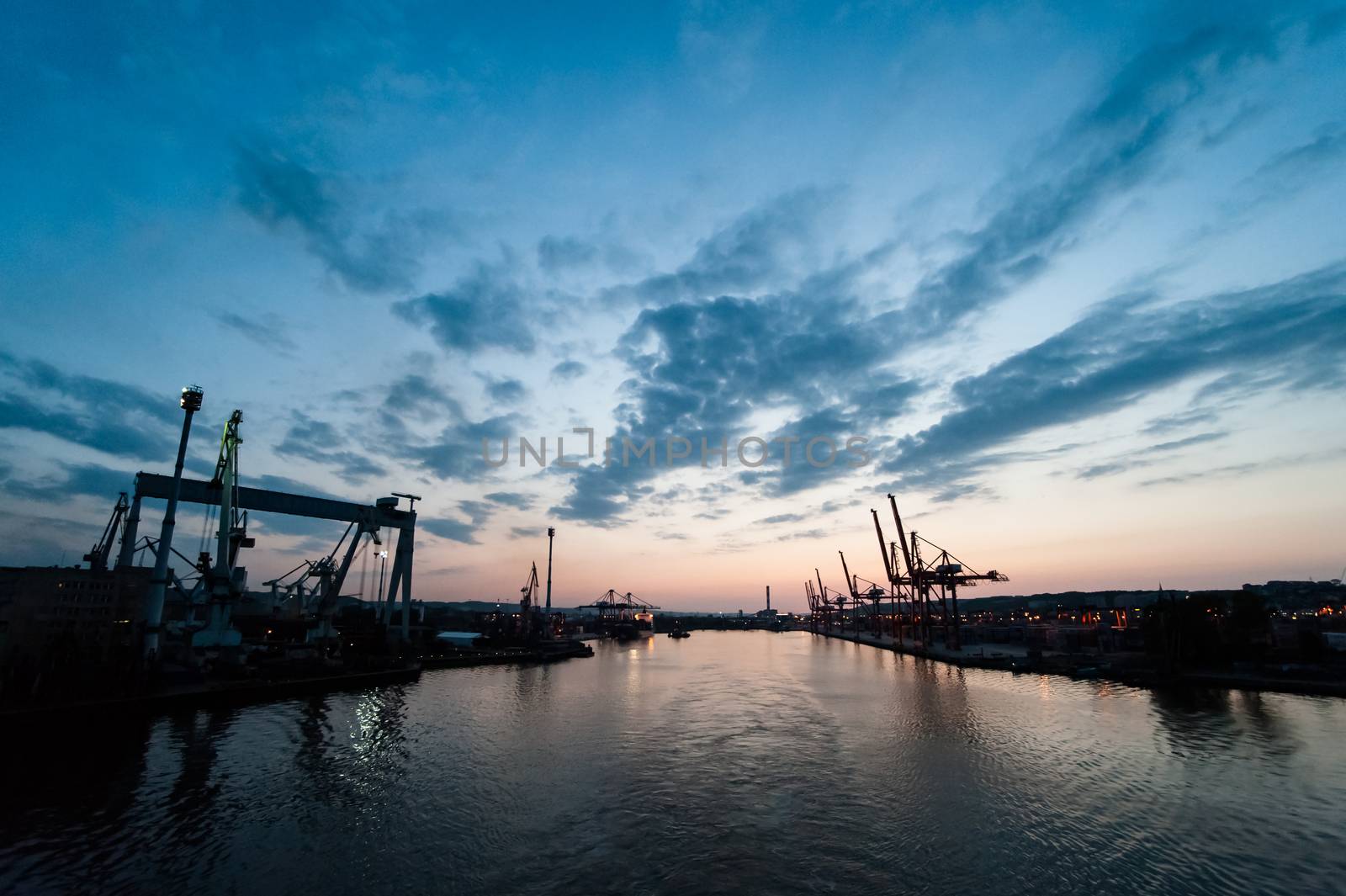 Dock cranes in cargo terminal with visible shapes of containers against dusk sky background, vivible port entrance and harbor.