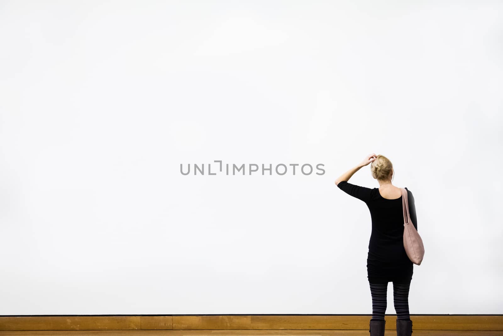 Caucasian Young Blond Woman Questioning in front of a Blank Wall in a Museum