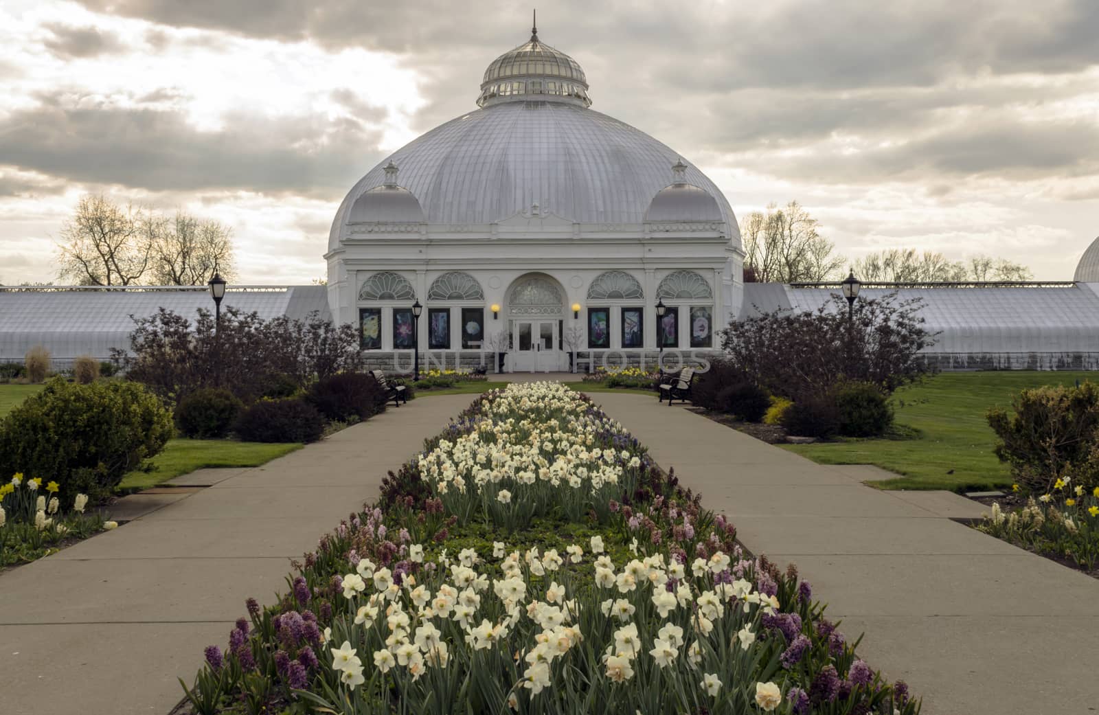 Rows of well maintained flower beds line the sidewalk leading up to an old domed and elaborate botanical garden structure. 