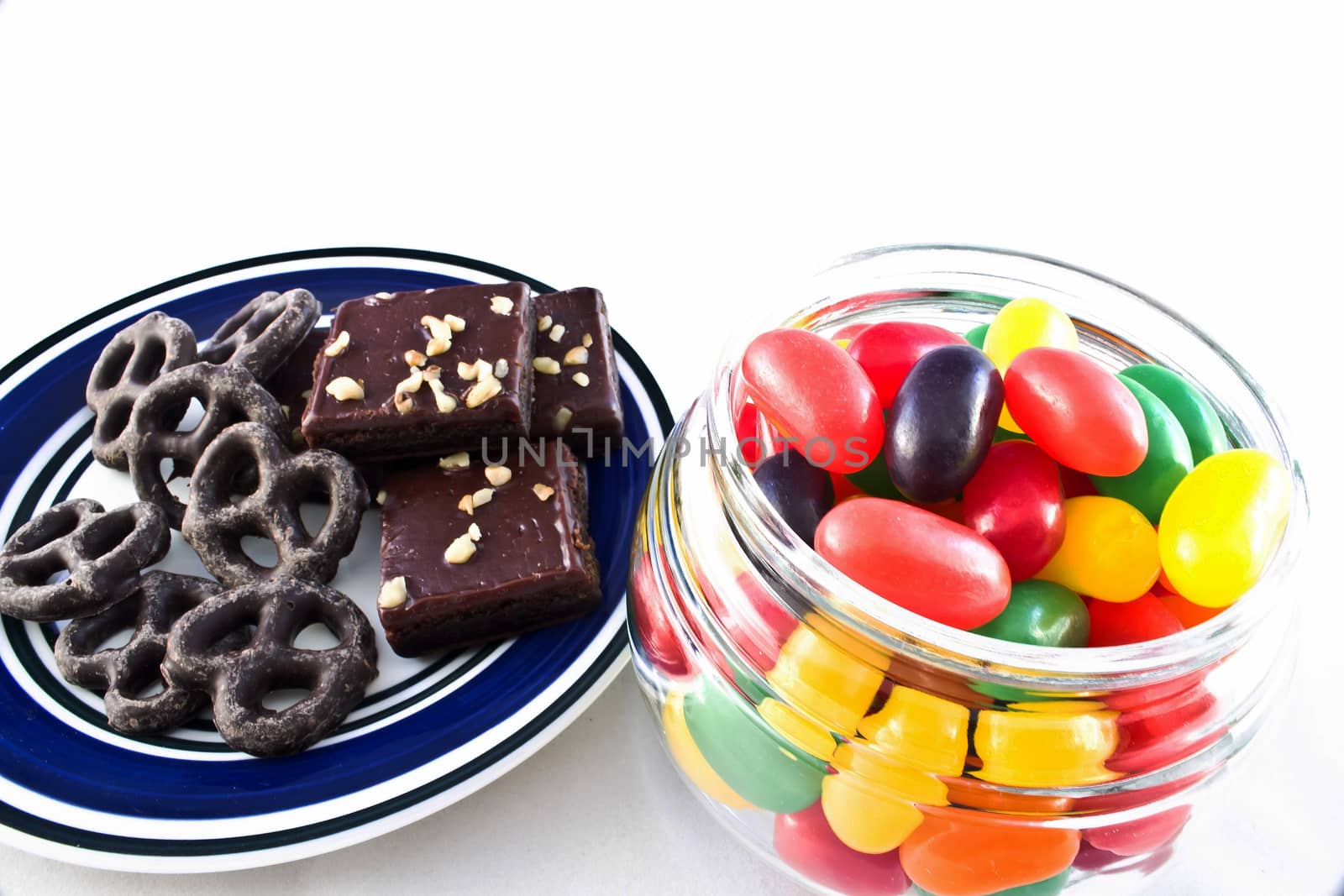 Plate of chocolate covered pretzels and walnut-covered fudge brownie squares. Jar of multi-colored jelly beans. 