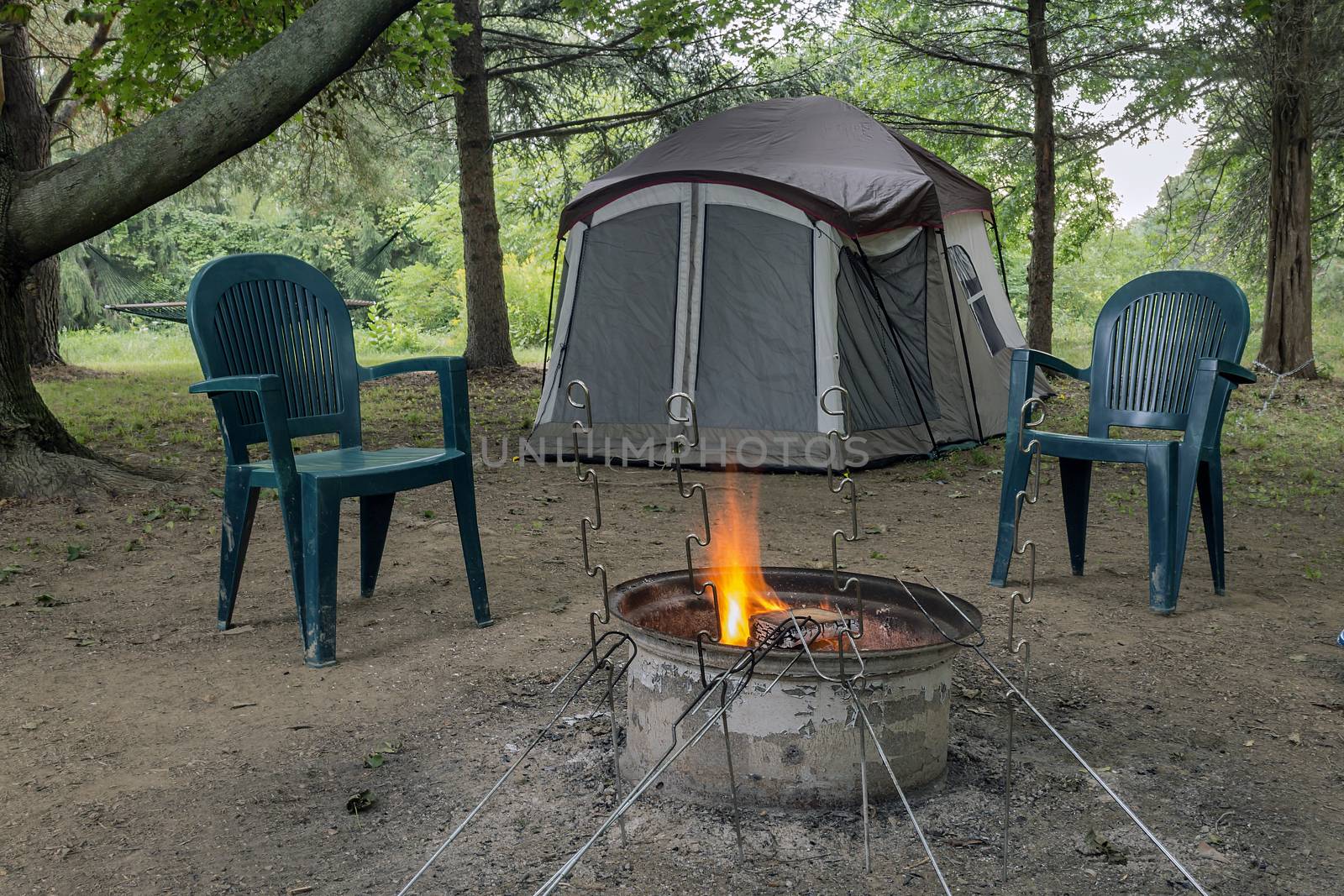 Pitched tent, crackling campfire, two chairs, and metal roasting sticks. All under a canopy of trees.