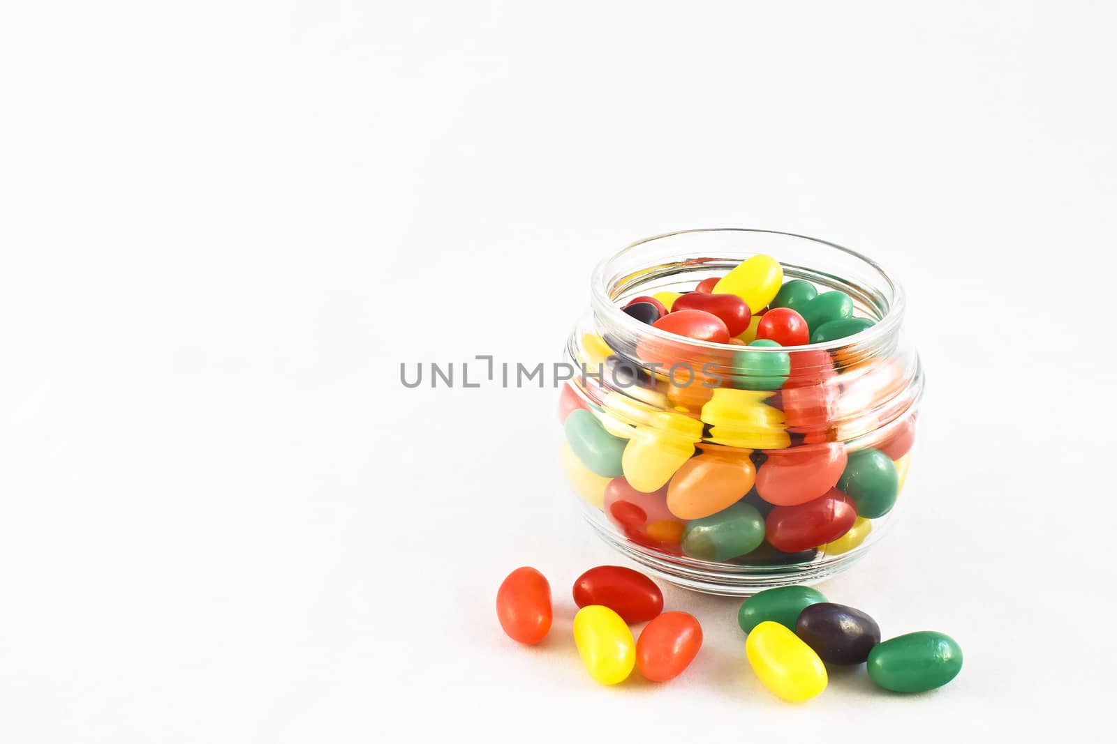 Glass jar full of multi-colored jelly beans. Several and strewn about on the white surface.