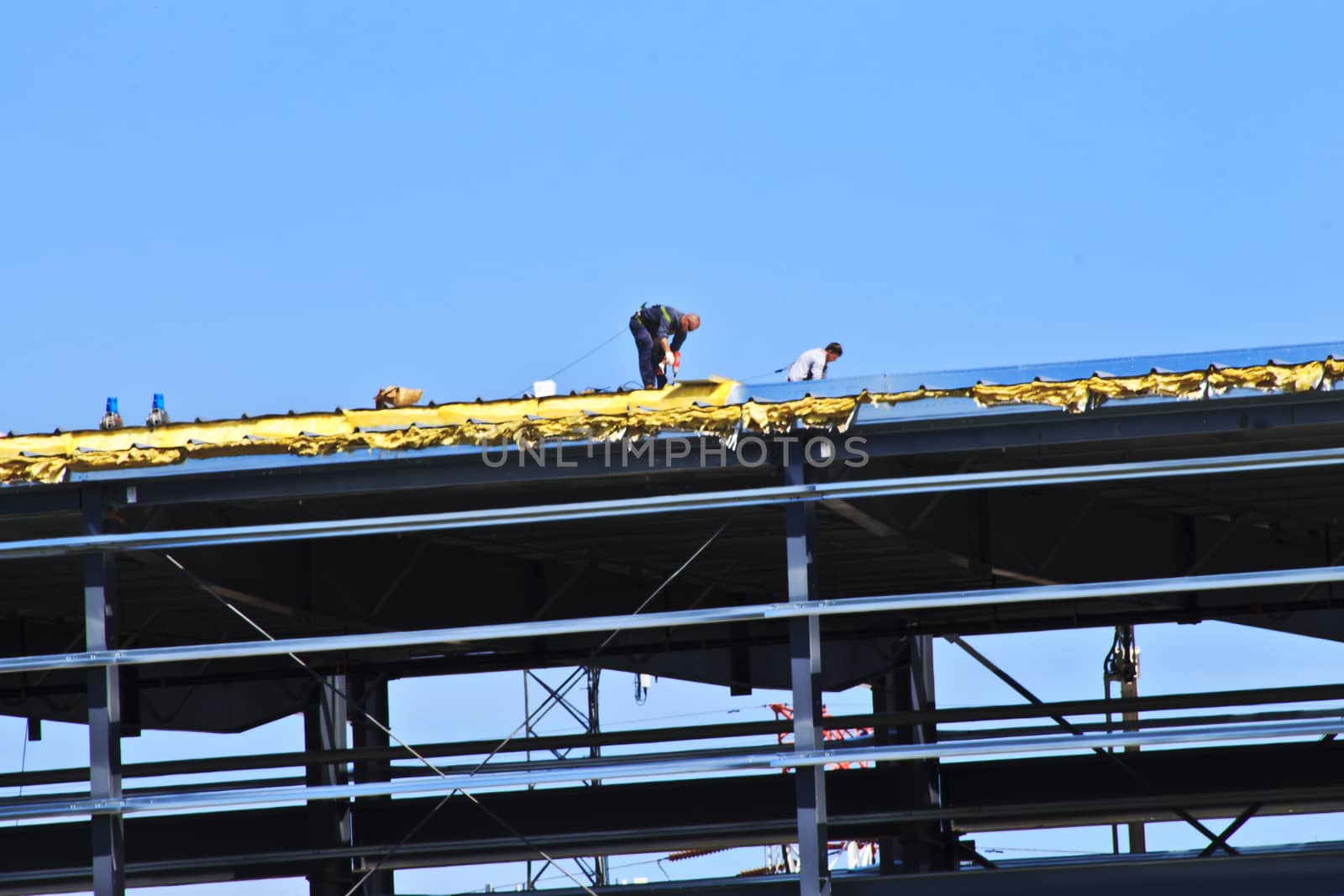 Two men are tethered to the roof of a high-story building. They are working on the roof of this new building under construction. 