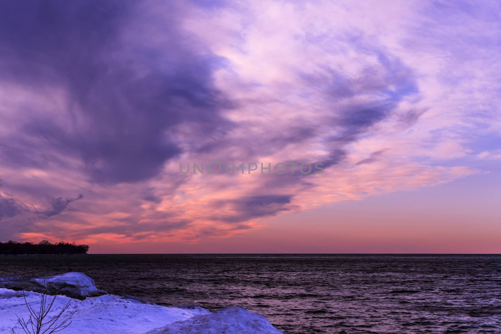 Lake with frozen shoreline and beautiful pink sunset with wispy purple hued clouds.