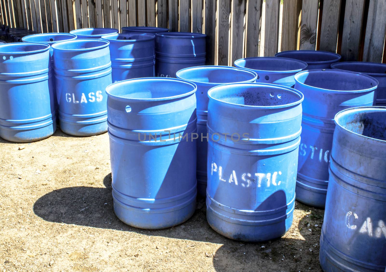 Many blue plastic barrels for recycling plastic, cans, and glass. They are inside a fenced area at a camping ground. 