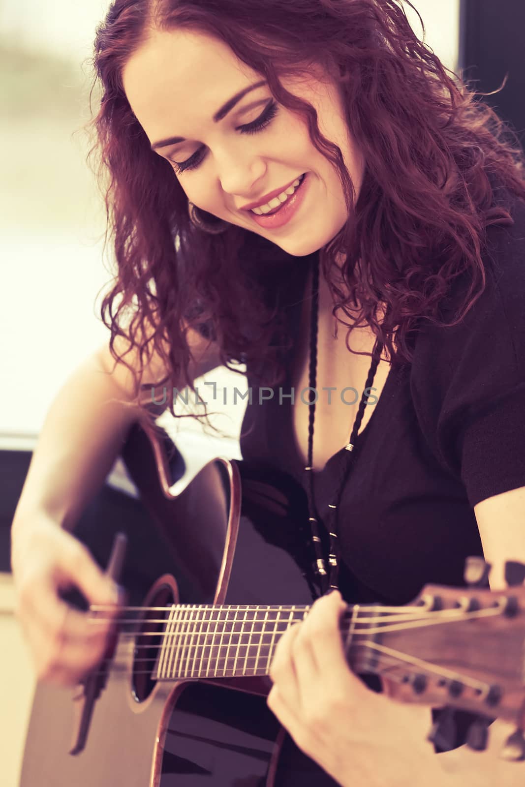 Photo of a woman playing an acoustic guitar by a window. Heavily filtered.

