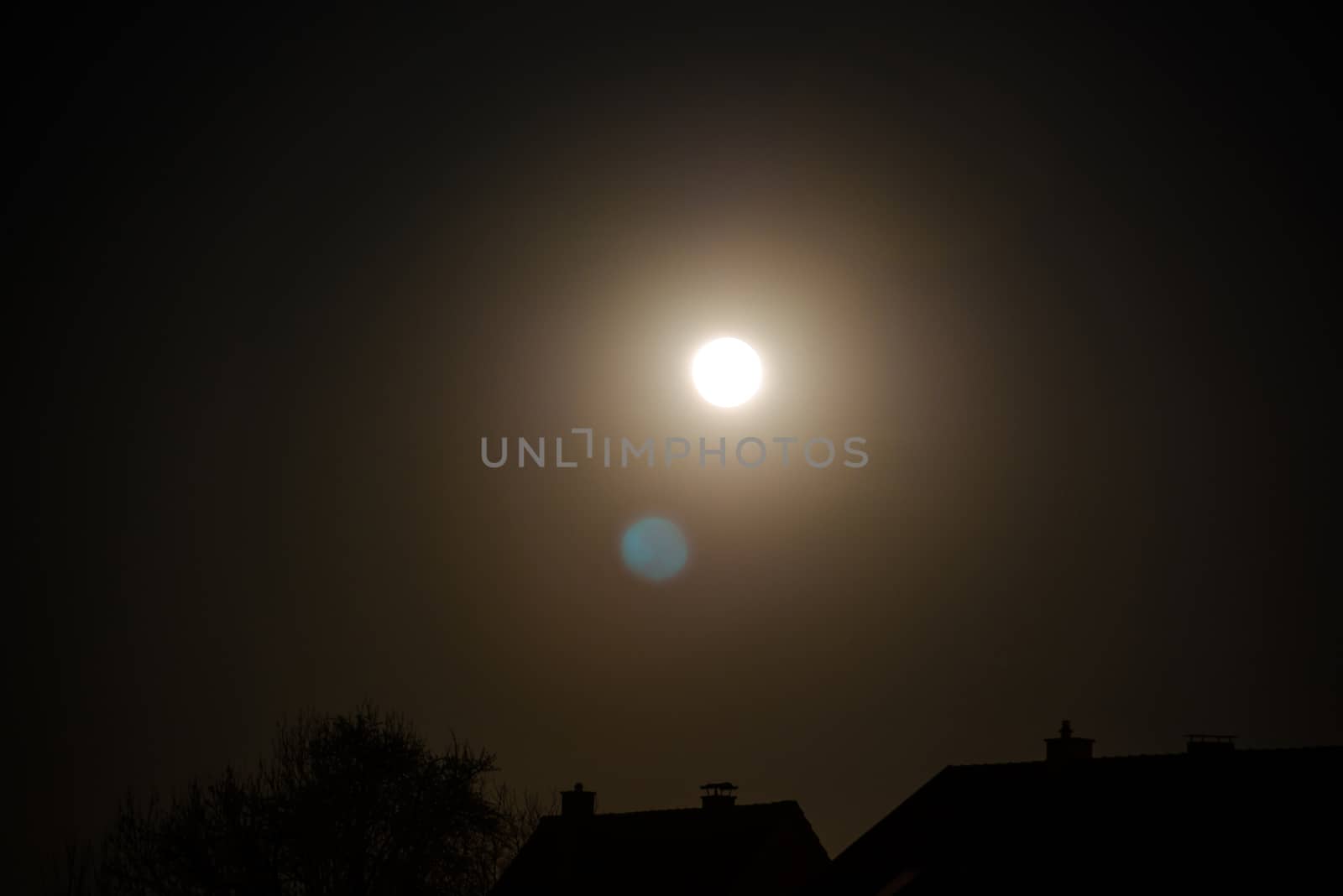 The moon being reflected by the camera lense and now visible in a second instance over buildings and trees