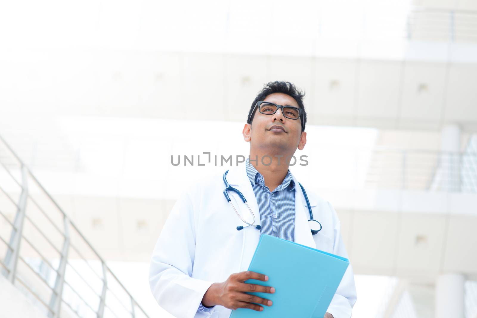 Portrait of an Asian Indian male medical doctor in lab uniform holding file folder standing in front of hospital.