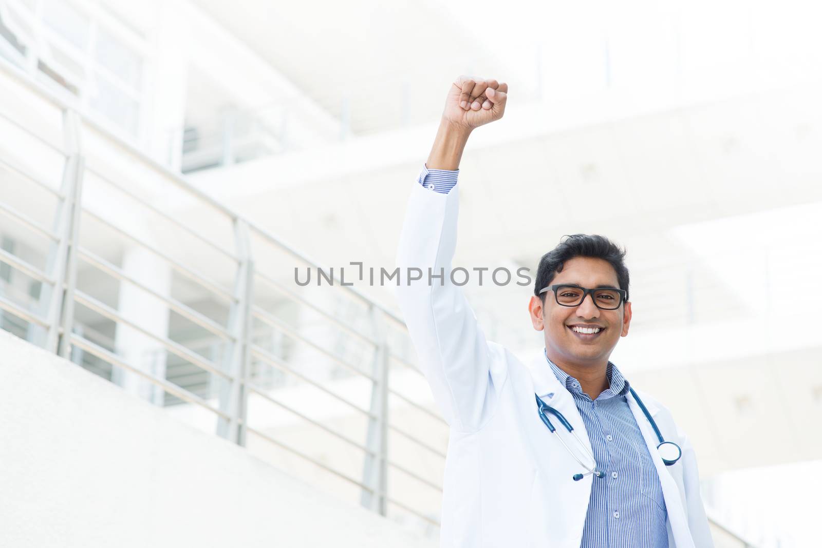 Successful Asian Indian medical doctor with arm up celebrating his victory, hospital building as background.