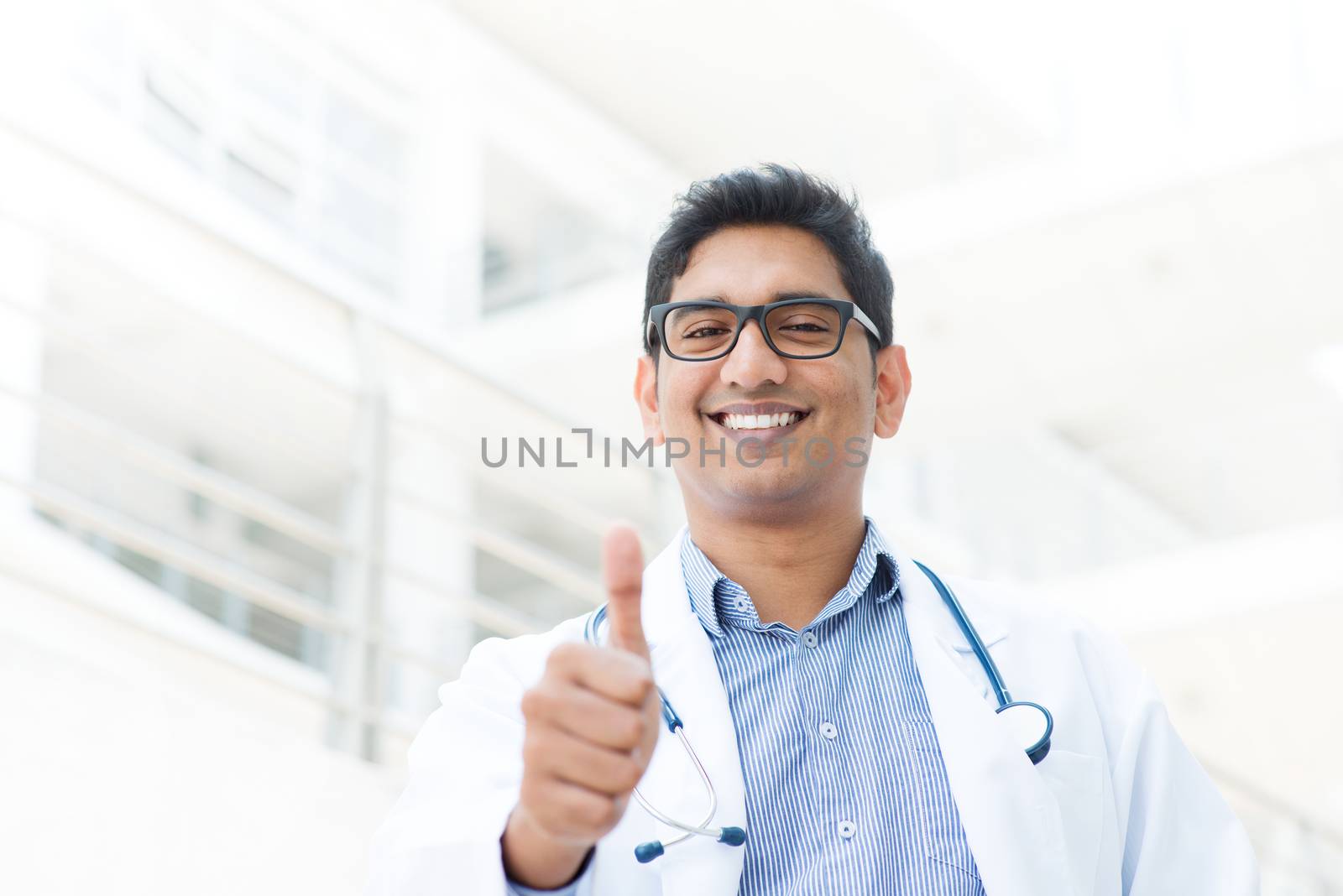 Portrait of a smiling Asian Indian male medical doctor in uniform thumb up, hospital building at background.