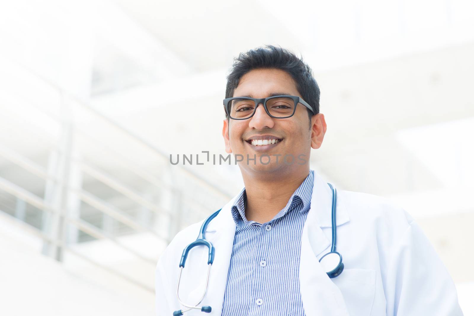 Portrait of a smiling Asian Indian male medical doctor in uniform, hospital building at background.
