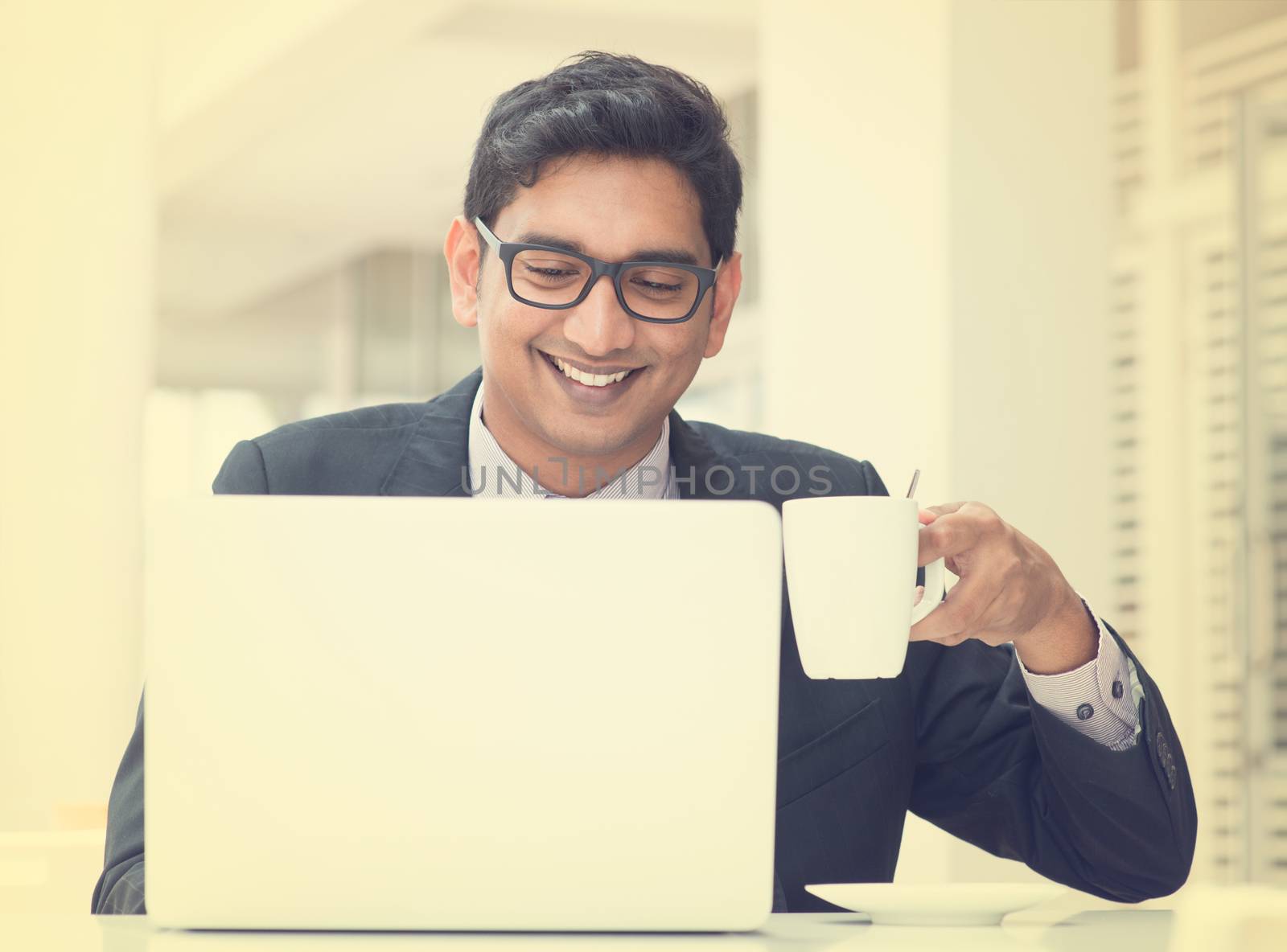 Young Asian Indian businessman using a notebook computer or laptop during office break at cafe, relaxing with a cup of coffee. India male business man, in vintage retro effect.
