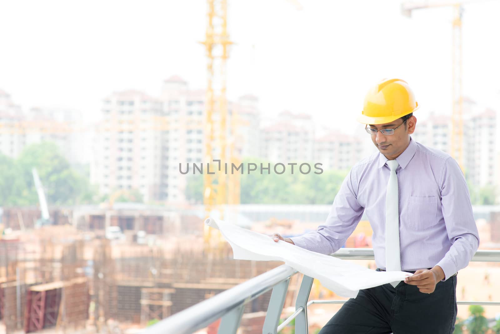 Portrait of a smiling Asian Indian male contractor engineer with hard hat reading blueprint at construction site.