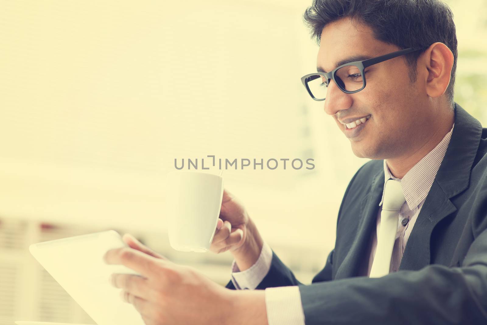 Young Asian Indian businessman using a tablet computer during office break at cafe, relaxing with a cup of coffee. India male business man,  in vintage retro style.