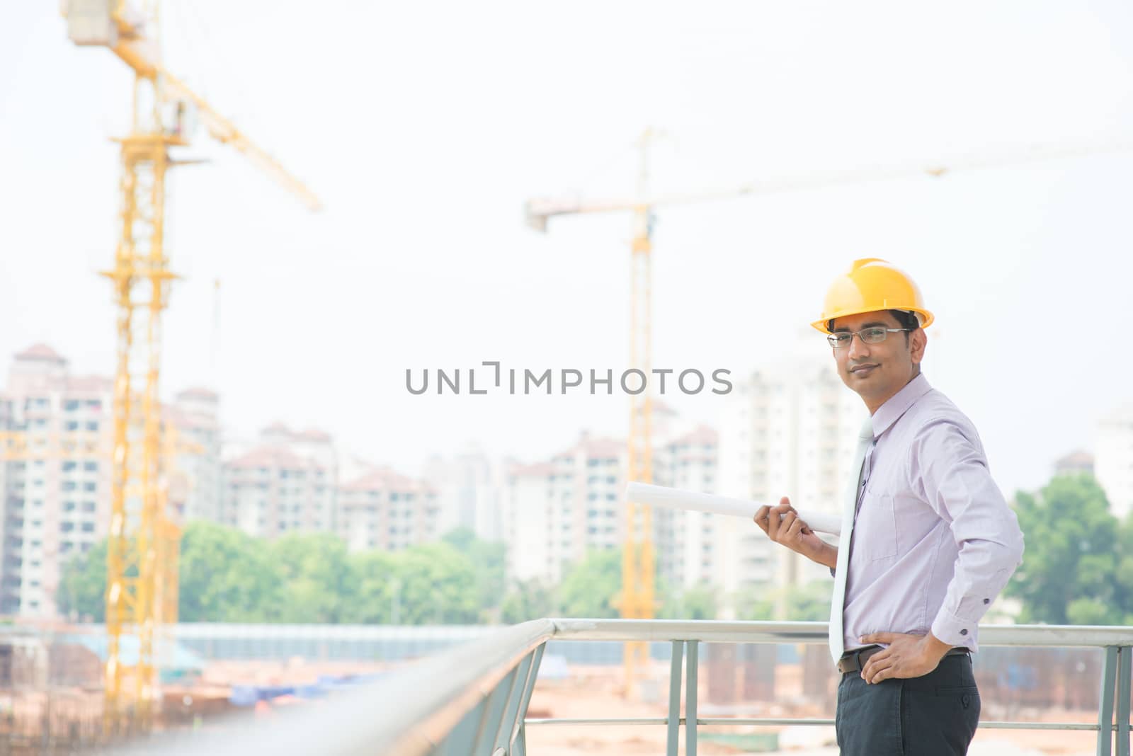 Portrait of a smiling Asian Indian male contractor engineer with hard hat standing at construction site.