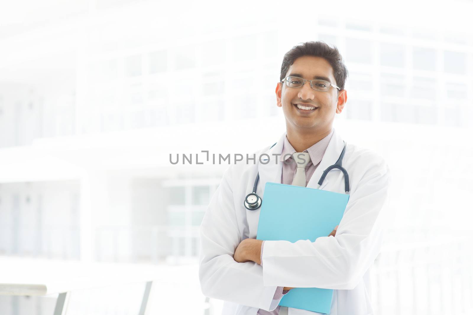 Portrait of a smiling Asian Indian male medical doctor standing inside hospital, holding file folder.
