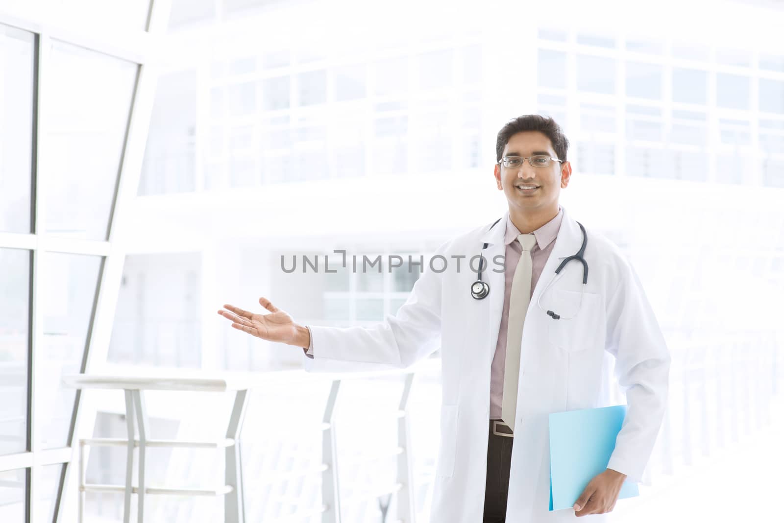 Portrait of a smiling Asian Indian male medical doctor standing inside hospital, holding file folder and showing welcome hand sign.