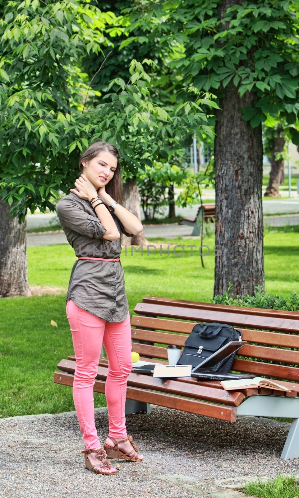 Young woman massaging her nape near a bench in a park during a short break of her work on laptop. 