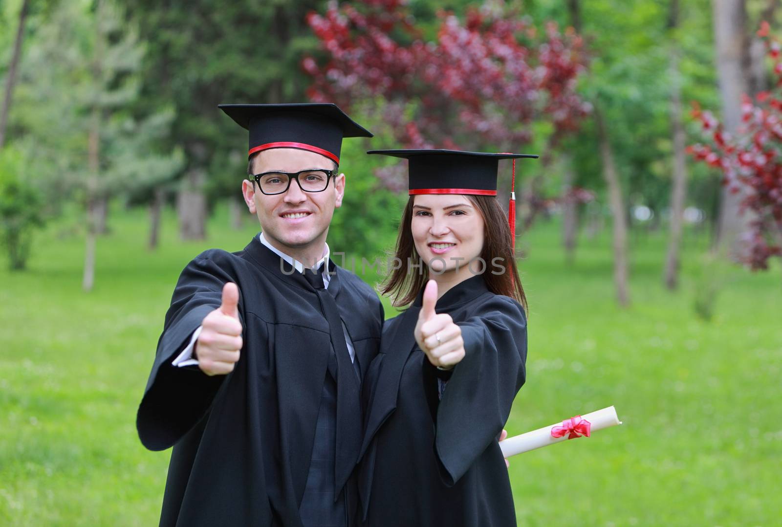 Portrait of a happy couple of students in the graduation day, posing with thumbs up in a park.