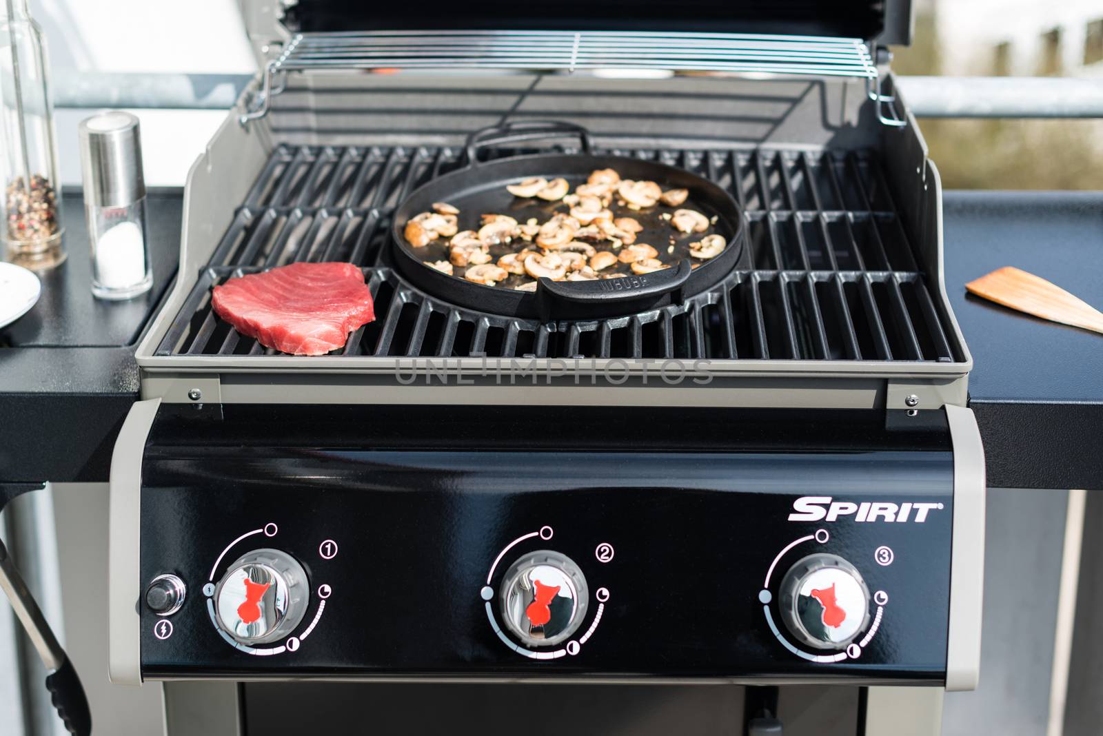 NEUHAUSEN, GERMANY - MARCH 27, 2014: A brand new Weber BBQ Gas Grill Spirit S320 black edition (model 2014) is waiting for its first use on March 27, 2014 in Neuhausen (near Stuttgart), Germany. The lid of the grill is open and reveals a lovely red tuna steak and some mushrooms being fried in the pan element in the center of the grill. Some utensils like BBQ tongs, a spatula, pepper and salt plus a plate are placed on its sideboards.