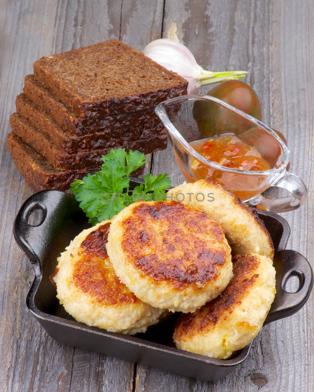 Arrangement of Delicious Chicken Meat Cutlets in Black Saucepan with Brown Bread, Bell Pepper Sauce and Vegetables closeup on Wooden background