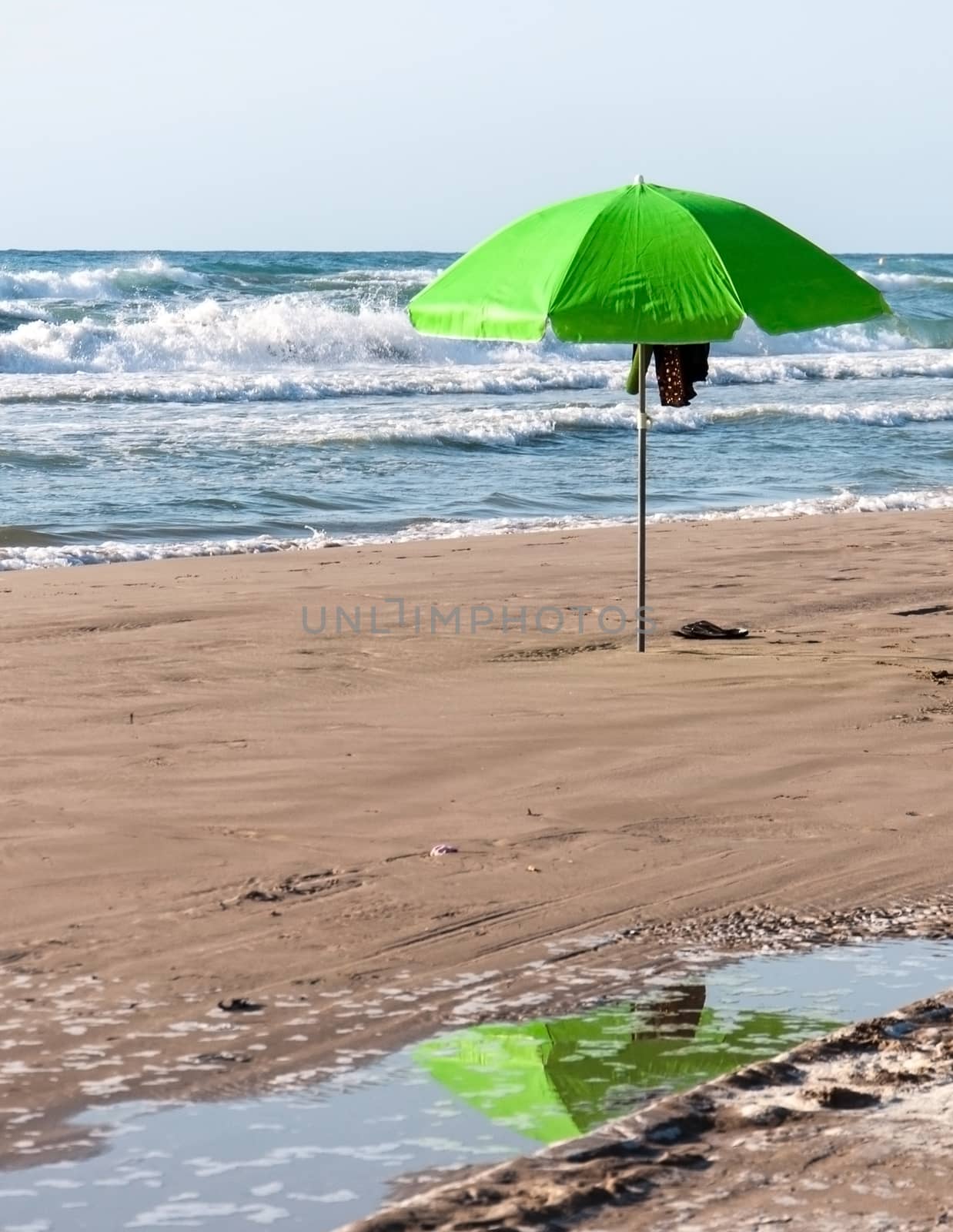 Green umbrella with reflection in pool of water on a beach.