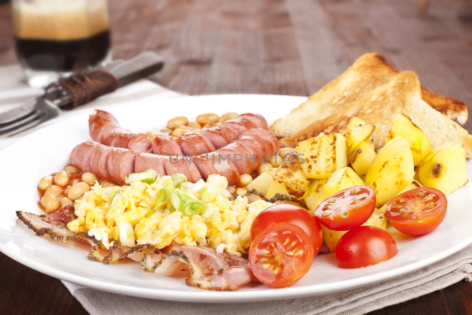 Delicious english breakfast, rustic style. Beans, sausages and scrambled eggs on white plate with fork. Coffee in background. Good morning concept.