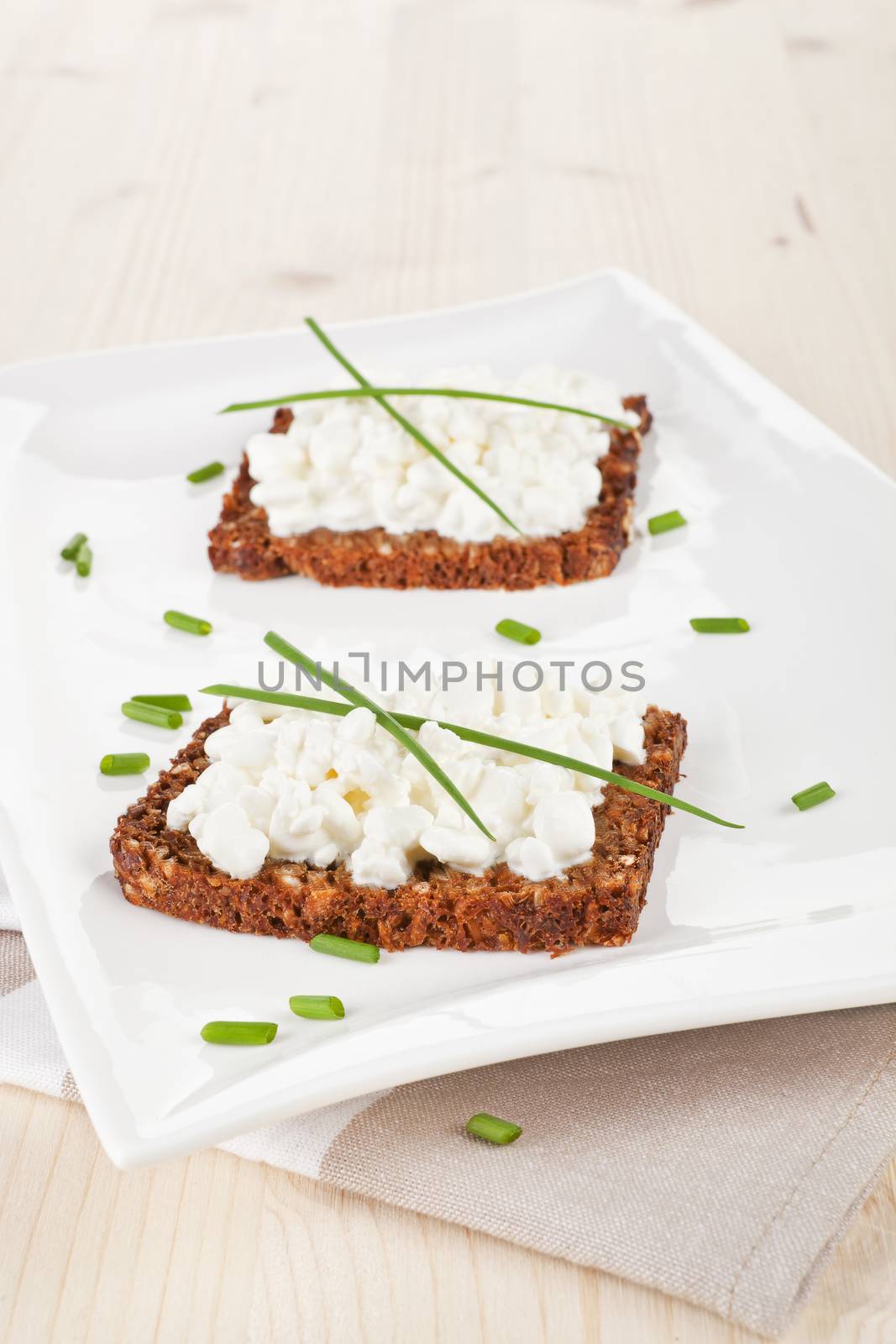 Delicious dark bread slices with cottage cheese and fresh herbs on white plate on wooden background.