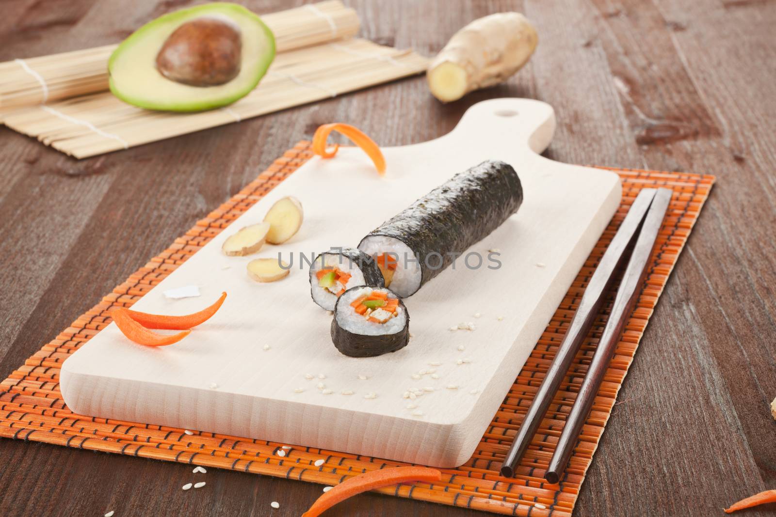 Delicious sushi roll on cutting board with bamboo mat, chopsticks, avocado and ginger.