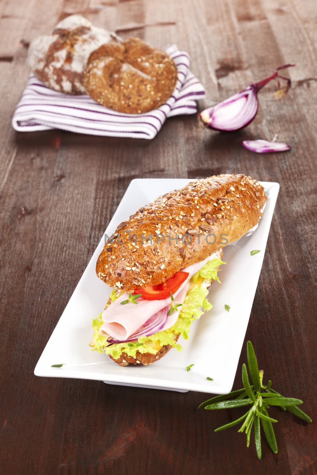 Wholegrain baguette with ham and fresh vegetables, decorated with onion and rosemary on brown wooden background. 