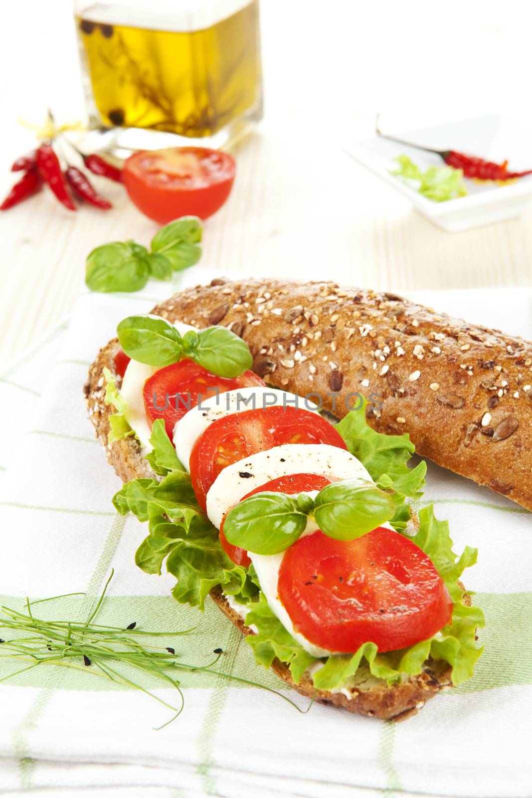 Mozzarella tomato baguette on wooden table, olive oil, herbs, tomatoes and chili peppers in background. Baguette caprese.